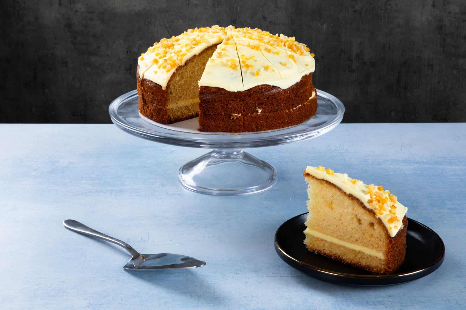 LHMC007-254.jpg Orange drizzle cake with creamy frosting and orange zest on a glass cake stand.