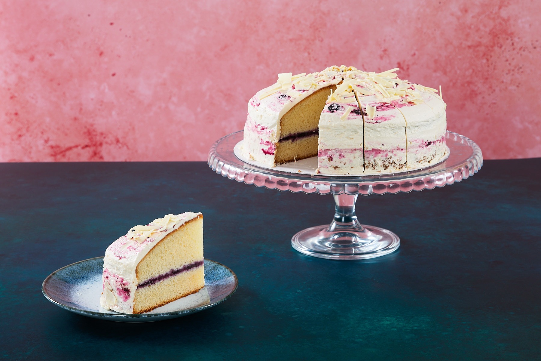 Lemon and Blueberry Lemon and blueberry cake with white chocolate topping on a glass cake stand.